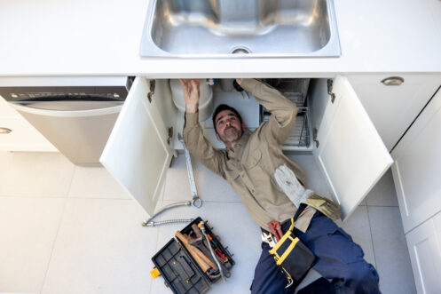 Plumber working under kitchen sink, tools and equipment visible, emphasizing professional plumbing services for home repairs.